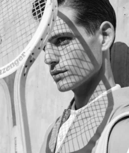Black and white close up of a man holding up a tennis racket and the sun creating shadows on his face.